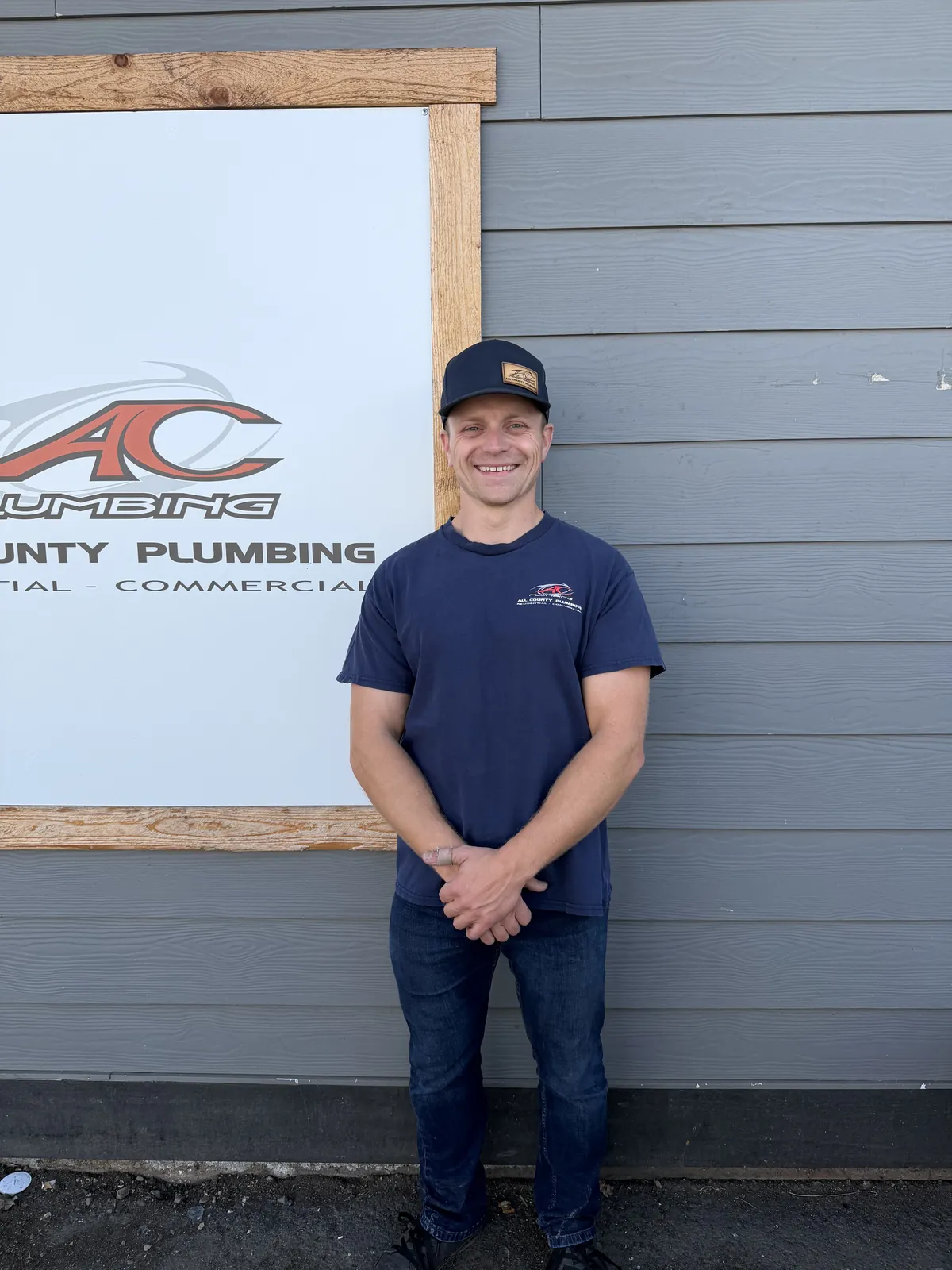 Josh in a navy All County shirt smiling in front of the AC Plumbing sign at the shop