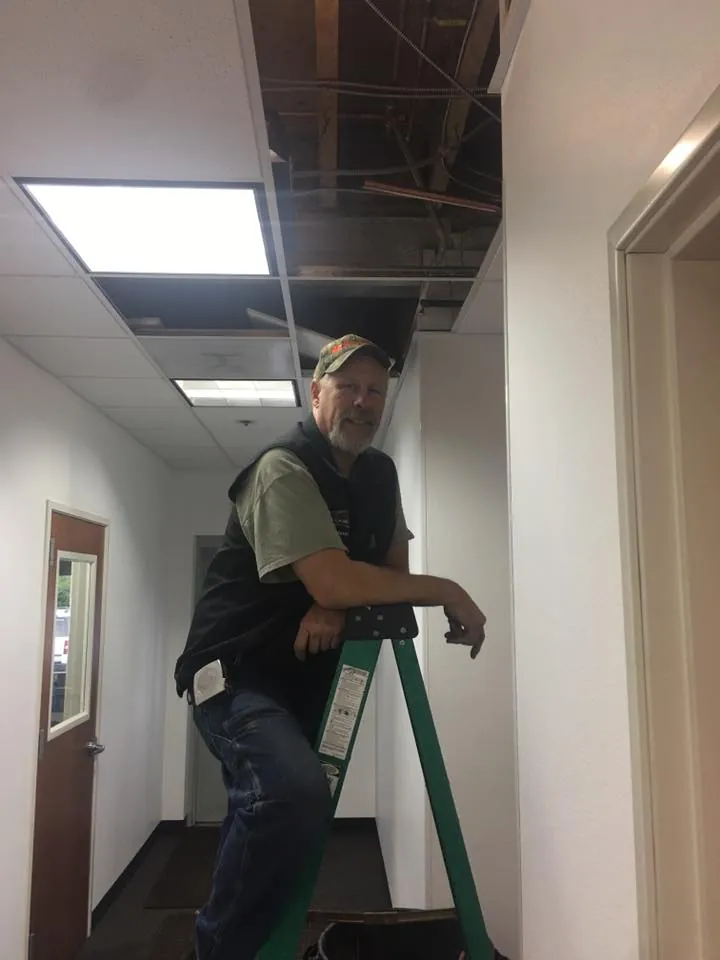 Plumber on a ladder below an open suspended ceiling in a commercial hallway